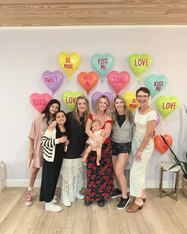 Friends and their kids gathered around a mother holding her baby in front of a wall of heart balloons