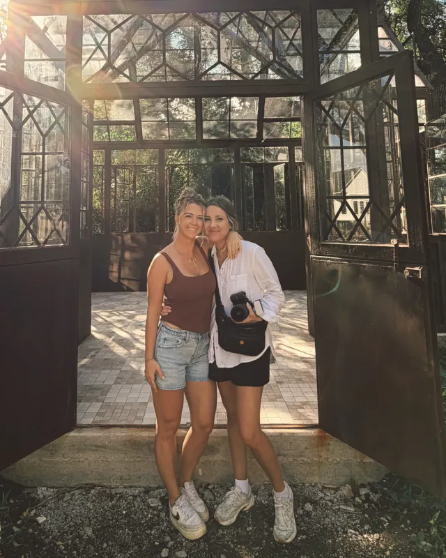 Two friends arm in arm in the open doorway of a glass garden house
